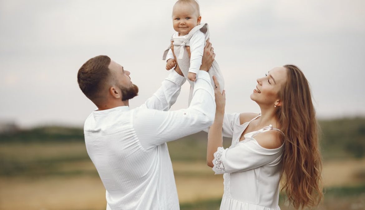 A joyful family scene with parents holding their smiling baby outdoors wearing white clothing.