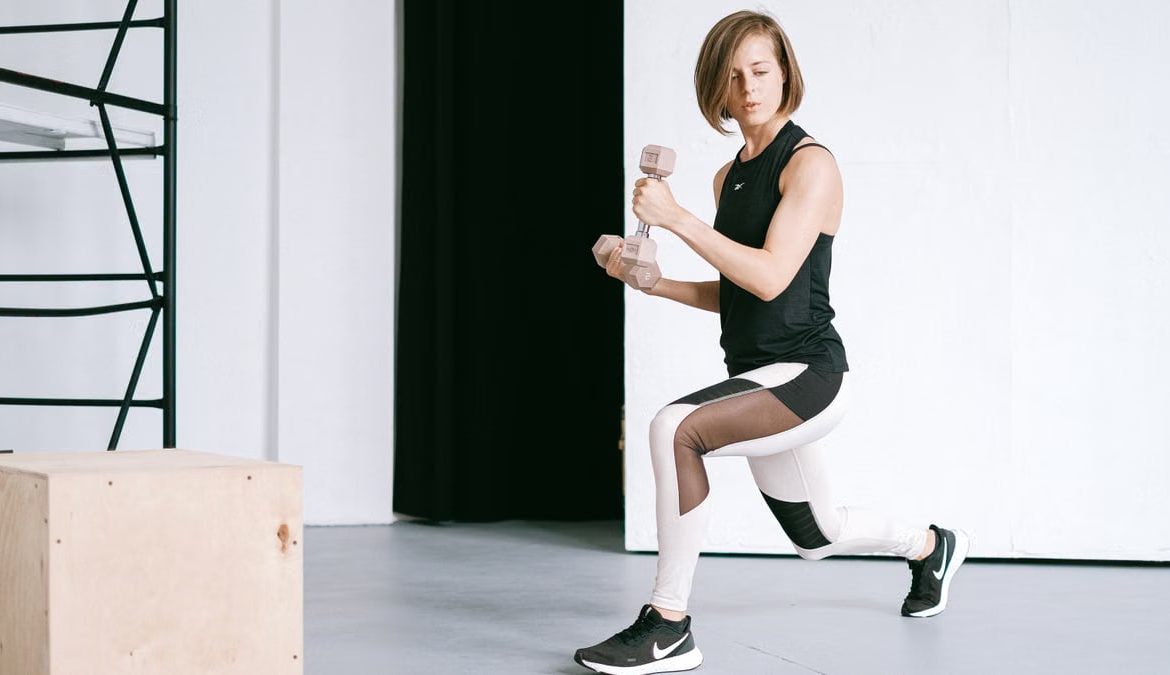 A young woman performing lunges with dumbbells in a modern indoor gym setting.